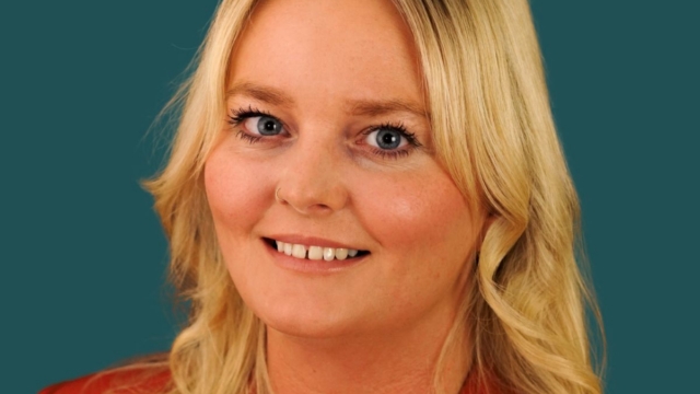 A professional headshot of a blonde woman wearing a red blazer and a delicate necklace, smiling warmly against a teal background.