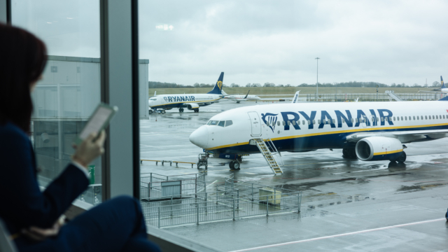Person viewing Ryanair aircraft on the tarmac from the airport terminal window, holding a mobile device and preparing for an upcoming flight, representing travel logistics and modern connectivity