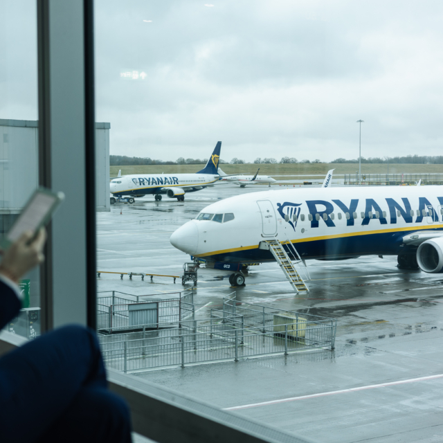 Person viewing Ryanair aircraft on the tarmac from the airport terminal window, holding a mobile device and preparing for an upcoming flight, representing travel logistics and modern connectivity
