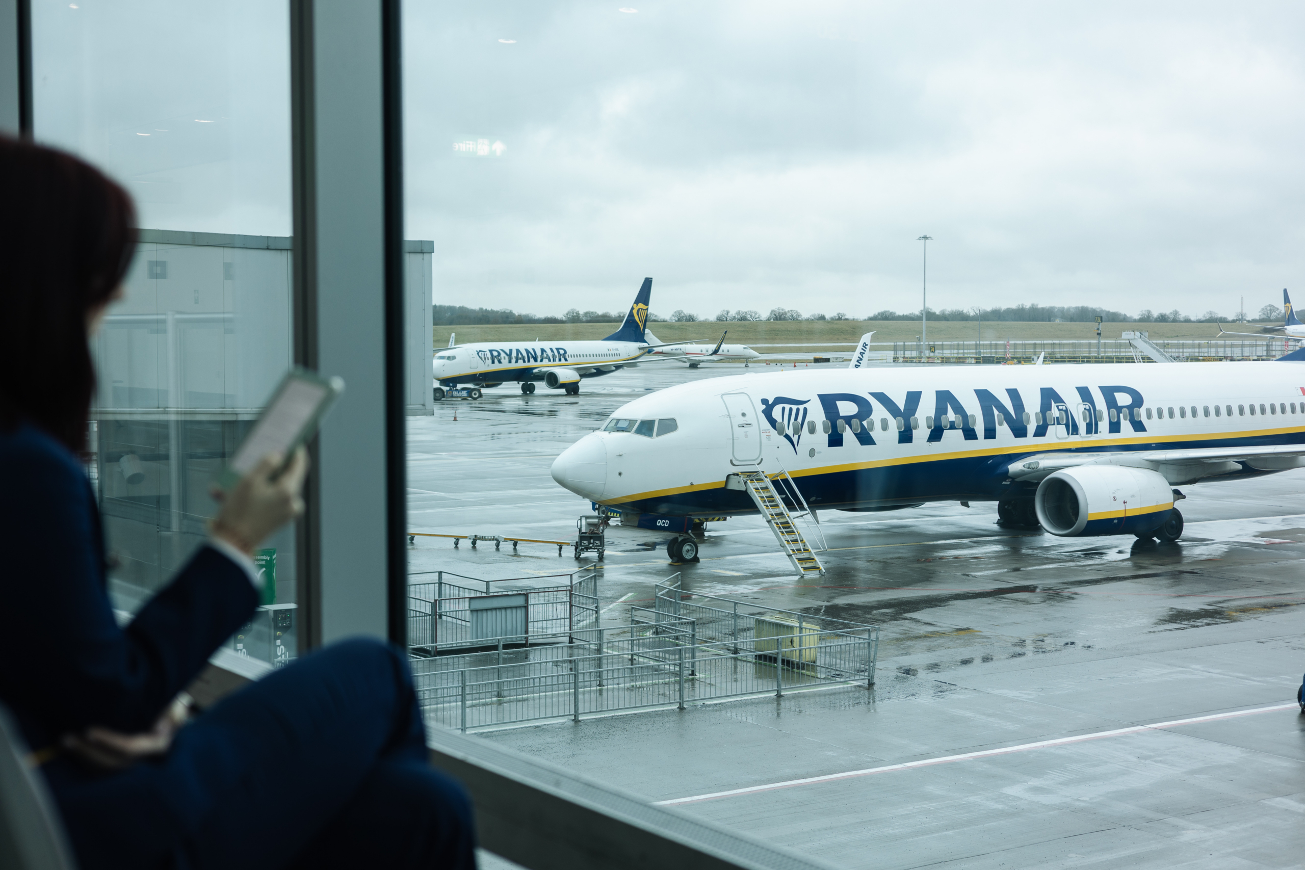 Person viewing Ryanair aircraft on the tarmac from the airport terminal window, holding a mobile device and preparing for an upcoming flight, representing travel logistics and modern connectivity